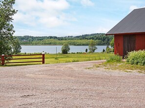 A gravel driveway leading to a red barn with a metal roof, surrounded by greenery and a lake in the background. - 6 Person Holiday Home in Odensvi (Gamleby)