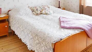 Una cama con cubierta de encaje blanco, cabecera de madera, mesita de noche con lámpara y papel tapiz floral.