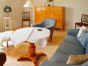 A room with a wooden floor, a grey armchair, a blue sofa, a white table with a white tablecloth, and a wooden cabinet. - 6 Person Holiday Home in Odensvi (Gamleby)