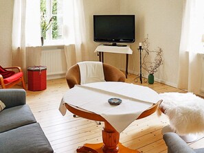 A living room with a wooden floor, a round table with a white tablecloth, a television on a stand, a red trash bin, and a grey sofa. - 6 Person Holiday Home in Odensvi (Gamleby)
