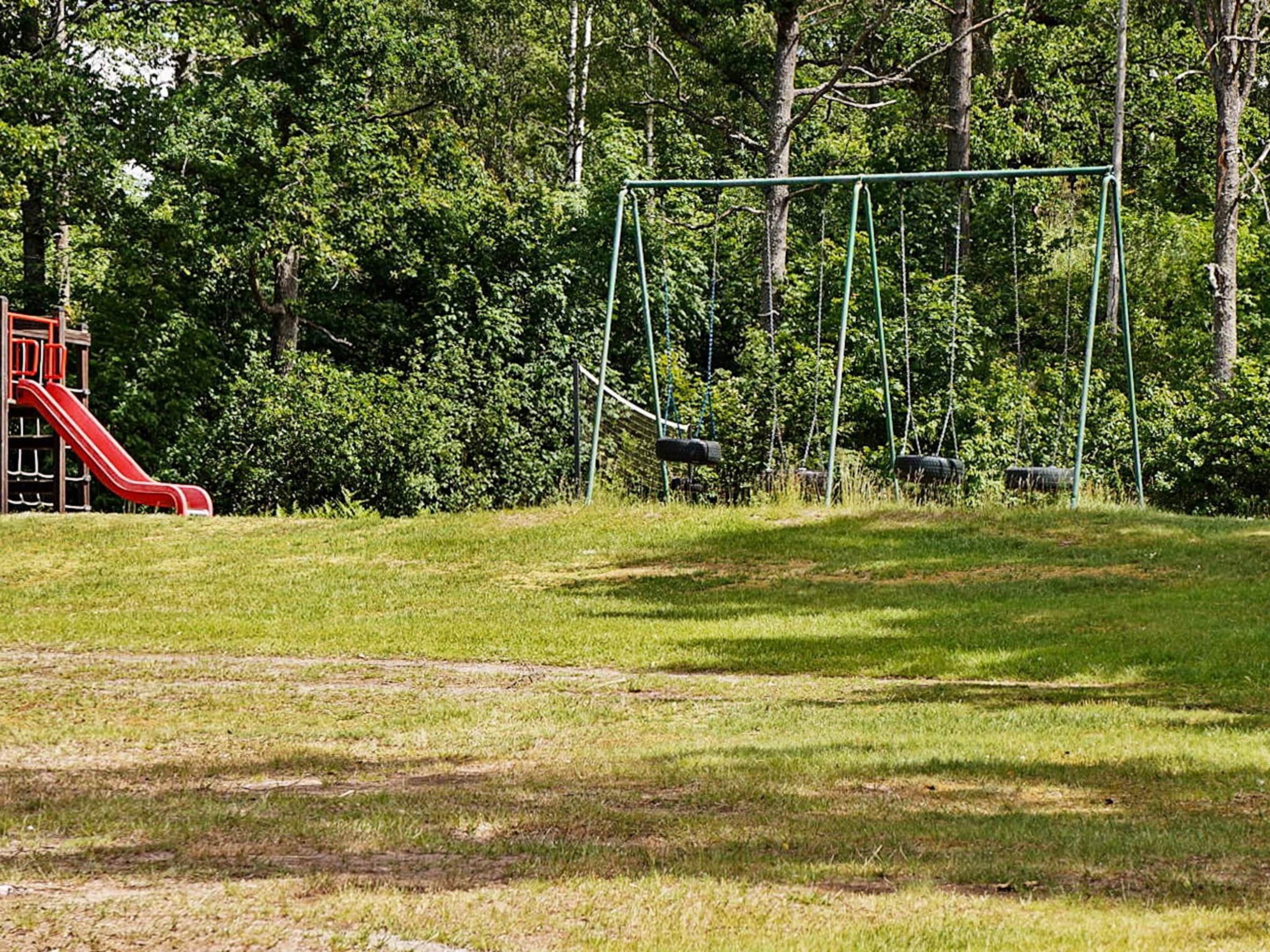 Un parque infantil con un tobogán rojo y columpios en un área verde con árboles al fondo.