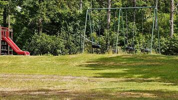 Un parque infantil con un tobogán rojo y columpios en un área verde con árboles al fondo.