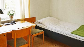 A room with a wooden desk, a chair, a bed with a white mattress and green blanket, and a window with curtains.