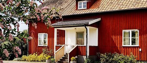 A red wooden house with a sloped roof, a front porch, and a small staircase leading to the entrance.