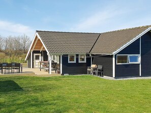 Une maison à deux étages avec un toit en pente, une véranda et une terrasse aménagée avec du mobilier d’extérieur.