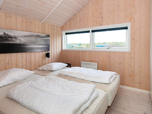 A room with wooden walls, a large window, two beds, a radiator, and a painting.