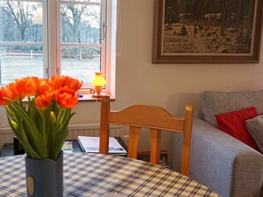 Ett rum med en trästol, ett bord täckt av en rutig duk, en vas med orange blommor, en lampa och en tavla på väggen.