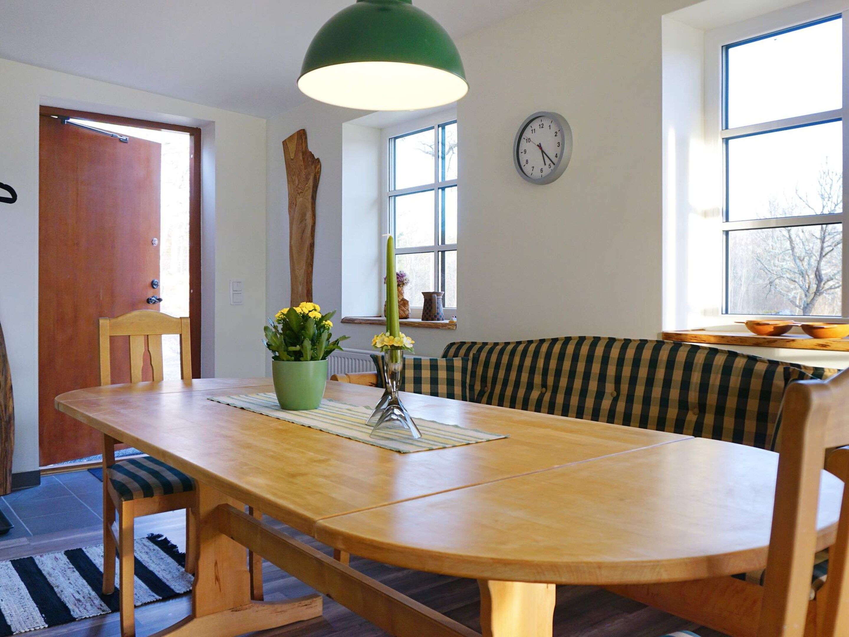 Un coin repas avec une table en bois, des chaises, un banc, une suspension verte, une horloge au mur et une fenêtre donnant sur un paysage agréable.