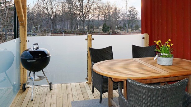 Une terrasse extérieure avec une table ronde en bois et quatre chaises en osier, un barbecue noir et un pot de fleurs orné de fleurs jaunes.