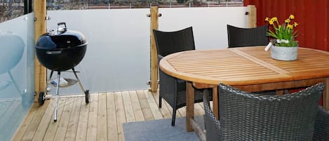 An outdoor patio with a round wooden table and four wicker chairs, a black grill, and a potted plant with yellow flowers.