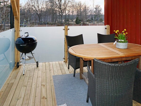 An outdoor patio with a round wooden table and four wicker chairs, a black grill, and a potted plant with yellow flowers.