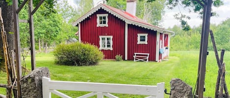 A red wooden house with a white gate in front, surrounded by greenery.