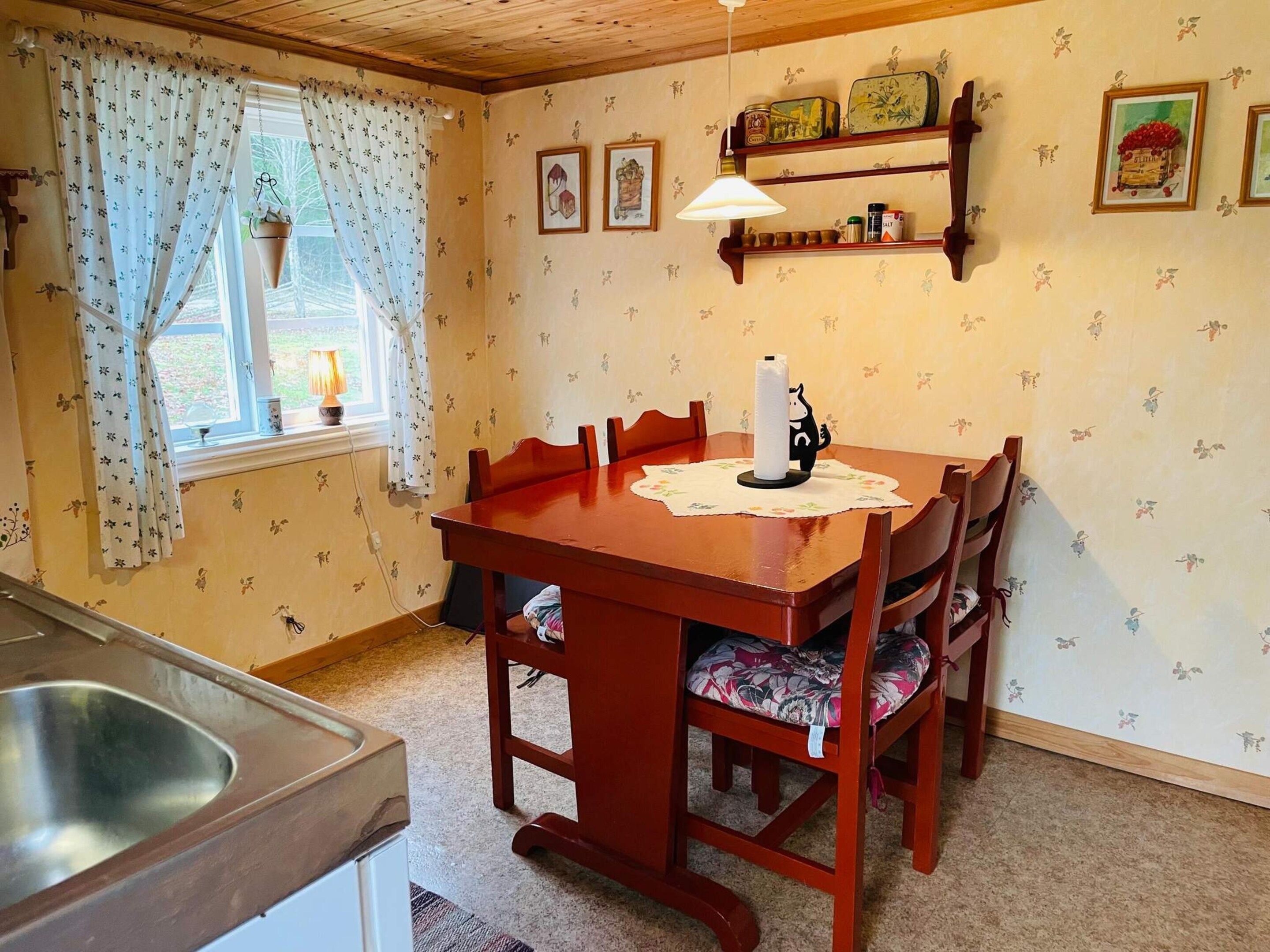 A dining area with a wooden table, chairs, a hanging light fixture, and a window with curtains.