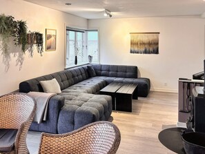 A modern living room with a sectional sofa, a dining table, wicker chairs, a large window with a view, and a wall-mounted plant.