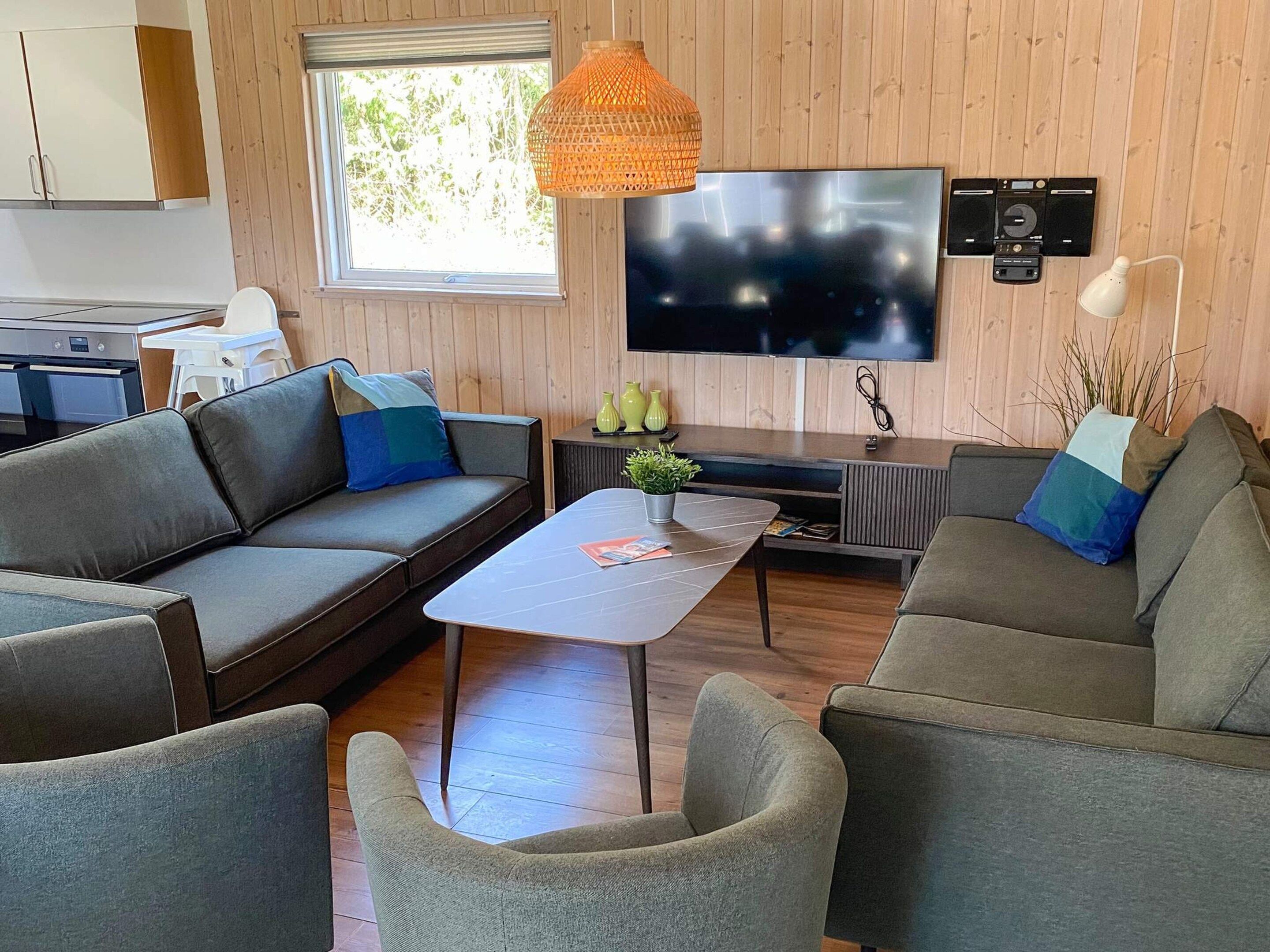 A modern living room with a sofa, armchairs, a coffee table, a TV, and a kitchen area.
