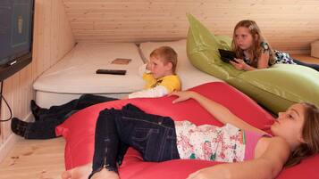 Ein Zimmer mit Holzboden, einem an der Wand befestigten Fernseher, einer Matratze und drei Kindern, die auf Sitzsäcken liegen.