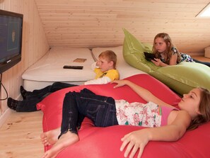 Une pièce avec un plafond en bois, un téléviseur fixé au mur, un matelas et trois enfants allongés sur des poufs.