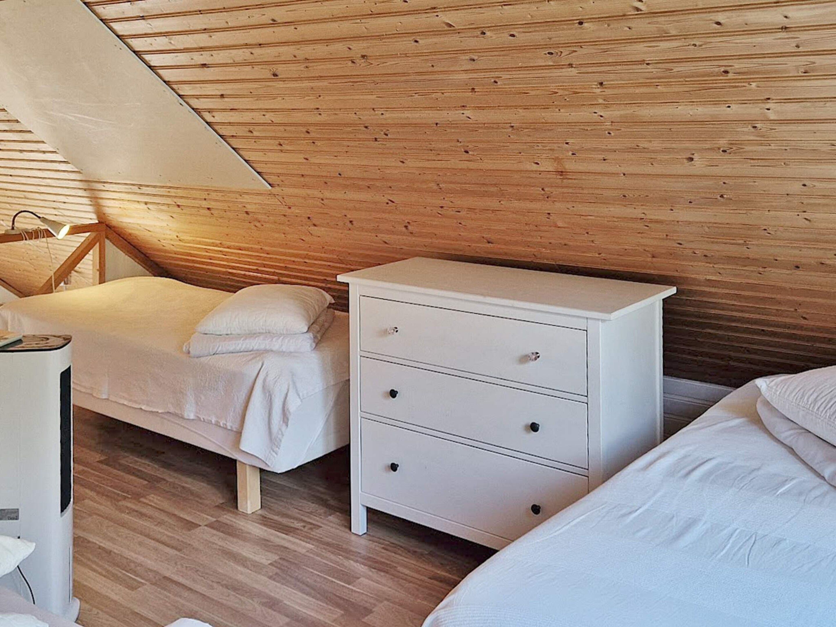 Une chambre avec un plafond en pente, des murs recouverts de lambris en bois, un lit, une table de chevet et une commode.