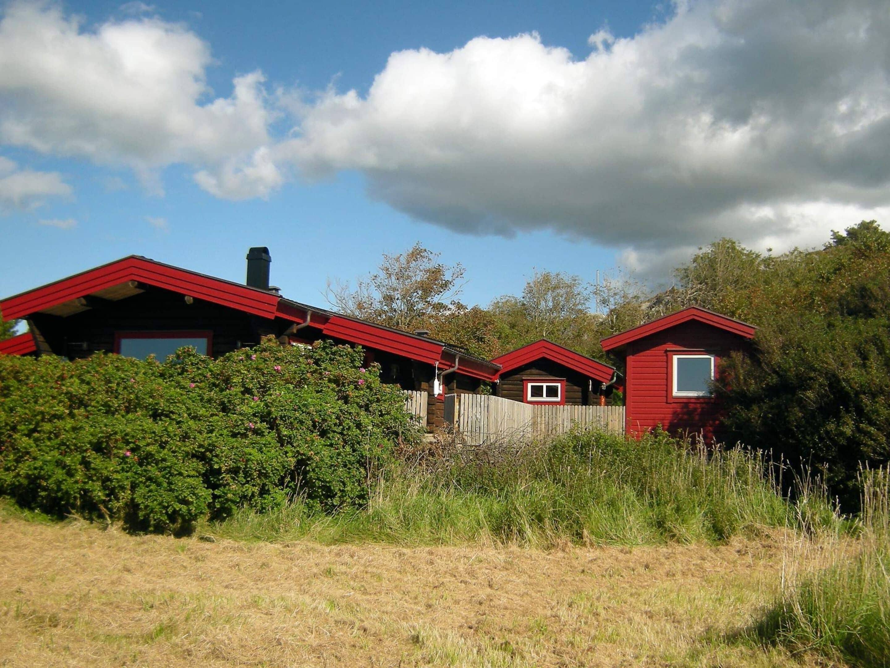 A row of red cabins with a wooden fence in front, surrounded by greenery and under a partly cloudy sky.