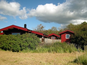 Une rangée de cabanes rouges, séparées par une clôture en bois, entourées de verdure et sous un ciel partiellement nuageux.
