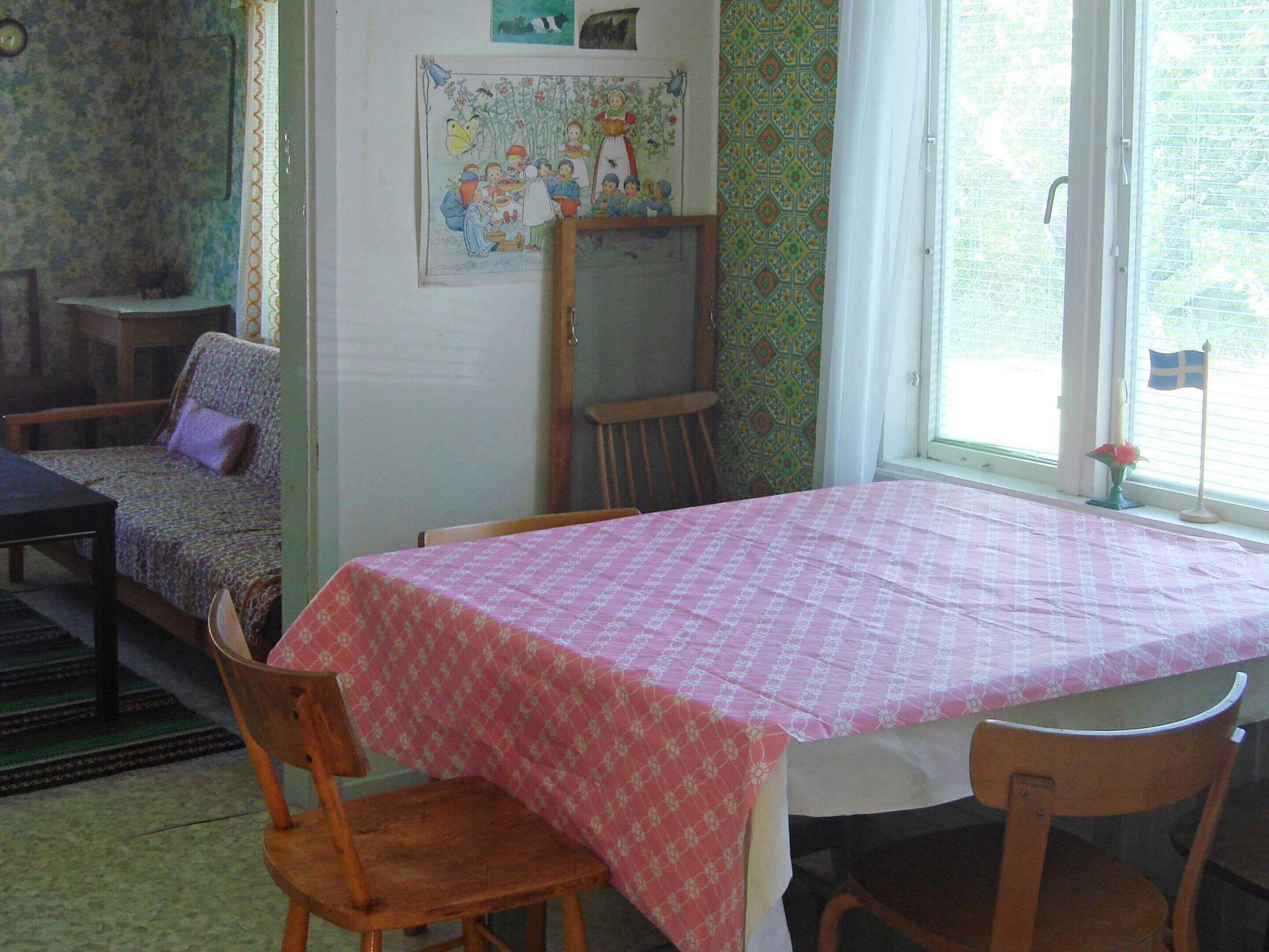 A room with a table covered in a patterned tablecloth, two wooden chairs, a sofa with a patterned cover, a wooden chair, a small table, and a window with a flag.