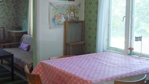 A room with a table covered in a patterned tablecloth, two wooden chairs, a sofa with a patterned cover, a wooden chair, a small table, and a window with a flag.