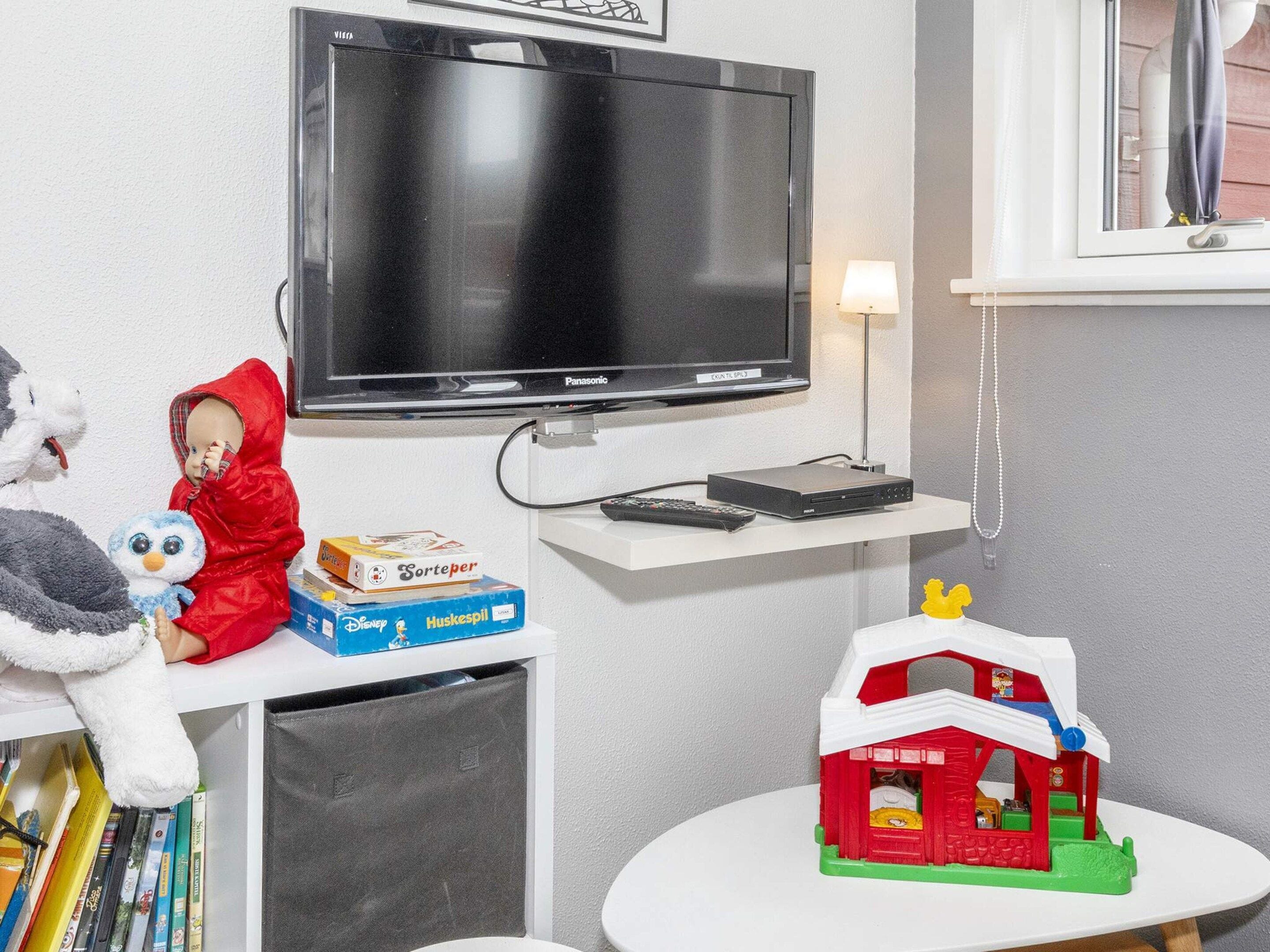 Et værelse med fjernsyn, et lille bord med et legetøjshus og en reol med bøger og en bamse.