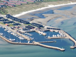 An aerial view of a coastal marina with boats docked, a pier, and a row of houses in the background.
