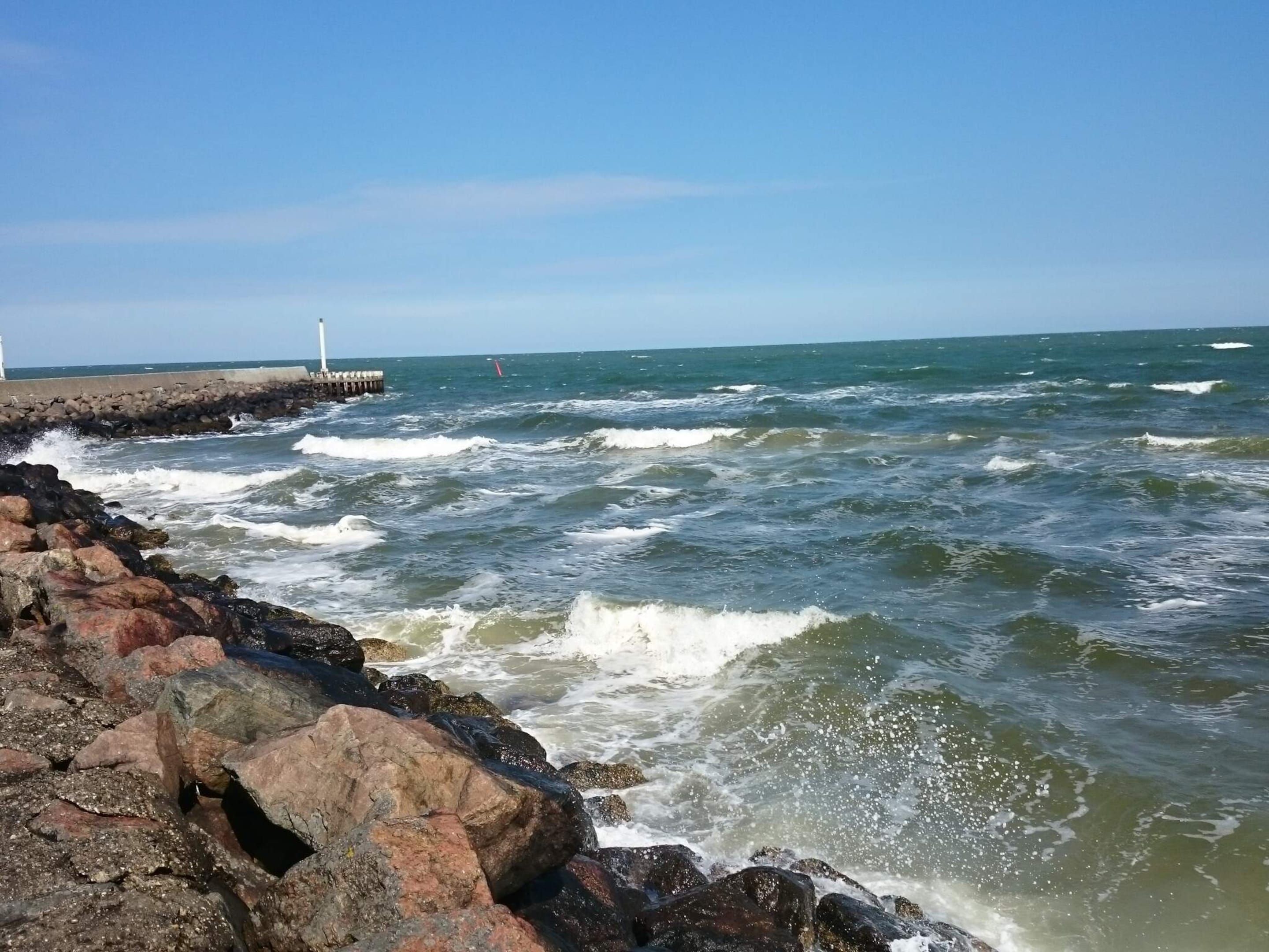 A rocky shoreline with waves crashing against it, a pier extending into the water, and a lighthouse in the distance.