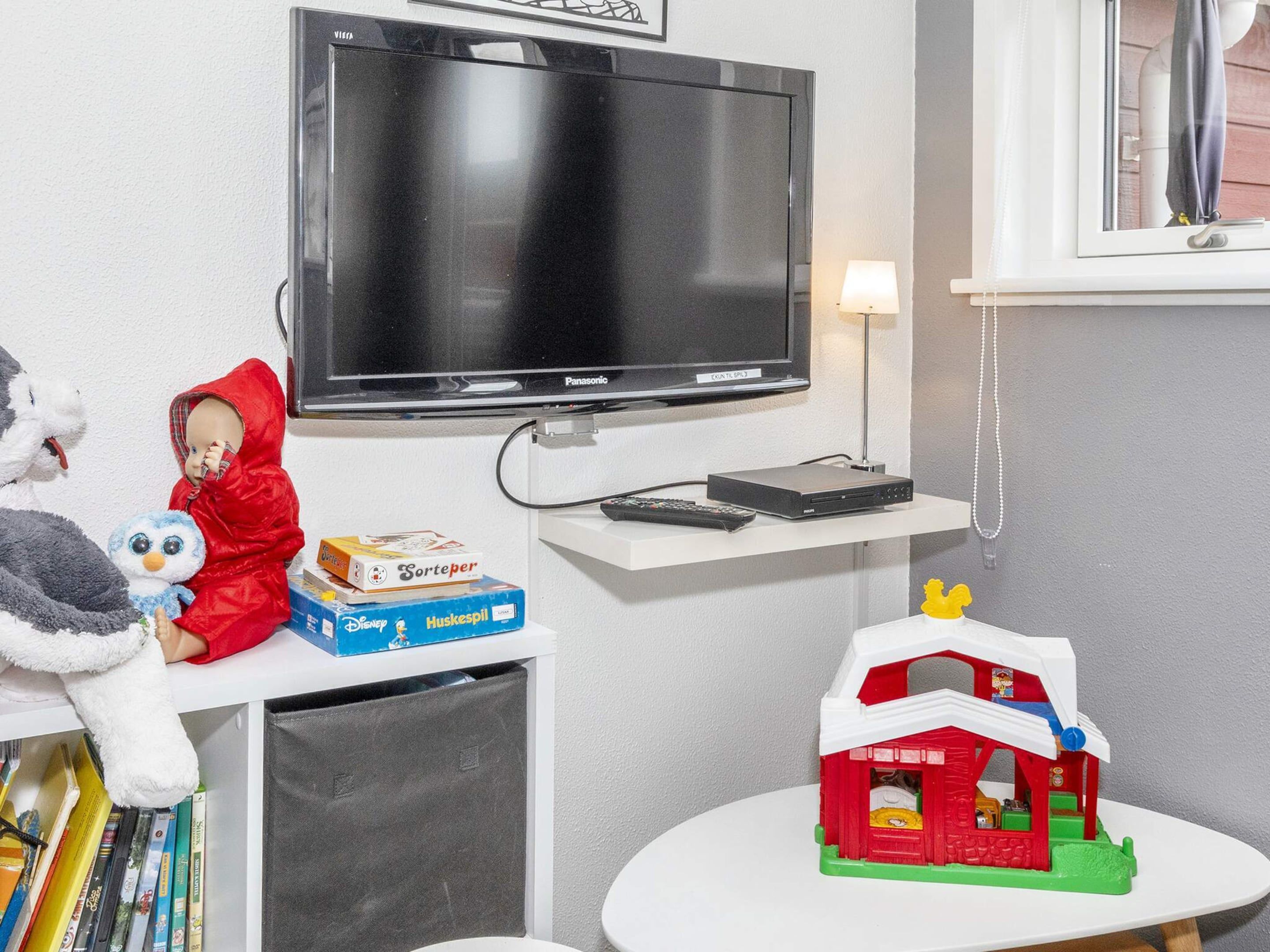 A room with a television mounted on the wall, a small table with a toy house, and a shelf with books and a stuffed toy.