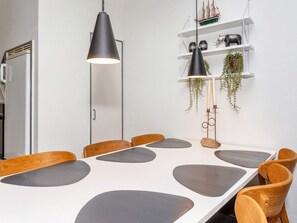 A modern dining area with a white table, grey placemats, wooden chairs, black pendant lights, and decorative items on shelves.