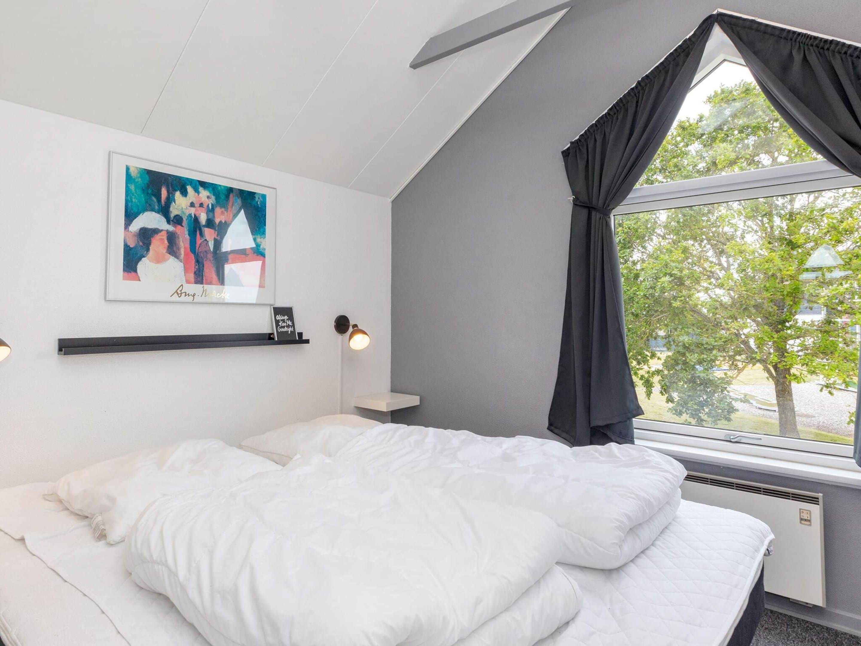 A bedroom with a bed, a window with curtains, a wall-mounted picture, and a bedside lamp.