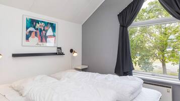 A bedroom with a bed, a window with curtains, a wall-mounted picture, and a bedside lamp.