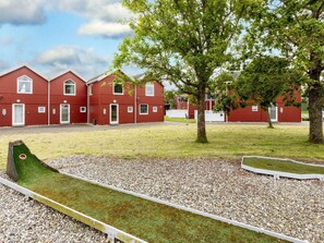 A red building with multiple windows and doors, surrounded by a grassy area with trees and a small green play structure. - Spacious Villa near Beach in Hadsund (Hadsund)