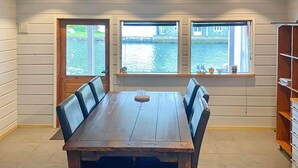 Un comedor con mesa y sillas de madera, ventanas amplias con vista a una masa de agua, y una estantería con objetos decorativos.