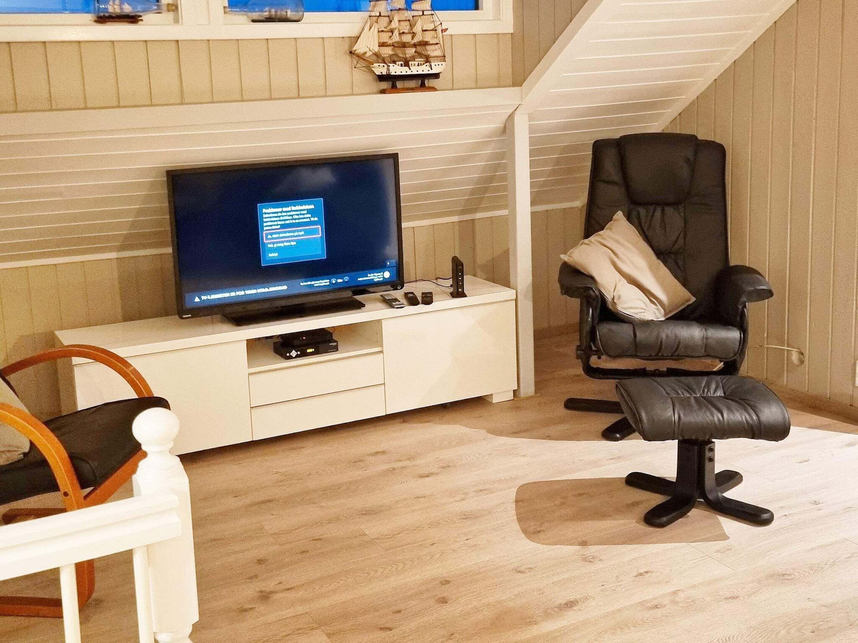 A living room with a television, a black office chair, a white chair, and a small ship model on the wall.