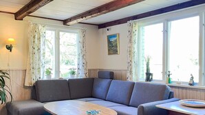 A cozy living room with a grey sectional sofa, a wooden coffee table, and a small wooden table with decorative items. There are two windows with curtains, a painting on the wall, and a lamp on the left. - 4 Person Holiday Home in Klippan (Klippan)