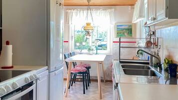 Una cocina con una mesa de comedor, sillas, un refrigerador, un horno, un lavaplatos y un ventanal con cortinas.