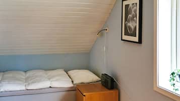 A small, single-bed room with a sloped ceiling, a bedside table, a framed picture on the wall, and a window with a view of greenery.