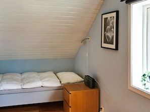 Habitació petita amb un llit individual, sostre inclinat, taula de nit, quadre amb marc a la paret i finestra amb vista a vegetació.