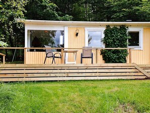 Una terraza de madera con una mesa, sillas, un ventanal y una puerta que da a un espacio interior.