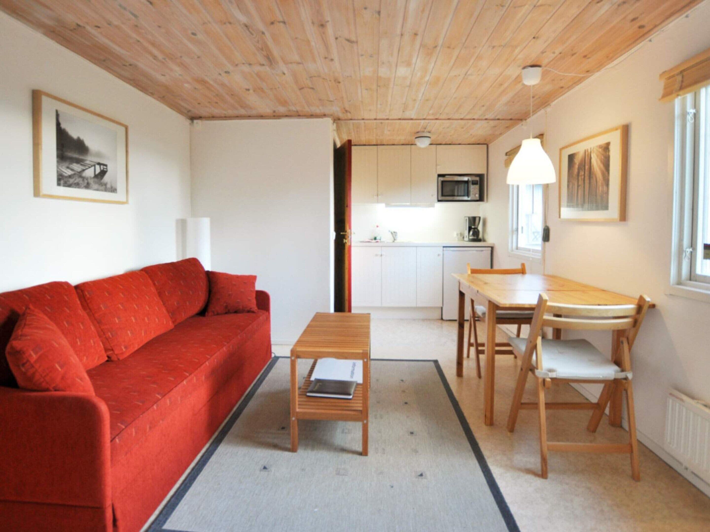 A compact living space with a red sofa, a wooden coffee table, a dining table with chairs, a kitchen area, and a framed picture on the wall.