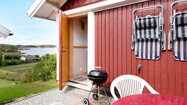 An open garage with a barbecue grill, two chairs, and a table outside a red building with a view of a lake. - 4 Person Holiday Home in Hamburgsund (Hamburgsund)