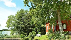 A red building with a white door, surrounded by lush greenery and a body of water. - 5 Person Holiday Home in Mariefred (Mariefred)