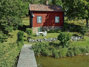 A red wooden house with a stone foundation, a small patio with a table and chairs, and a wooden dock leading to a body of water.