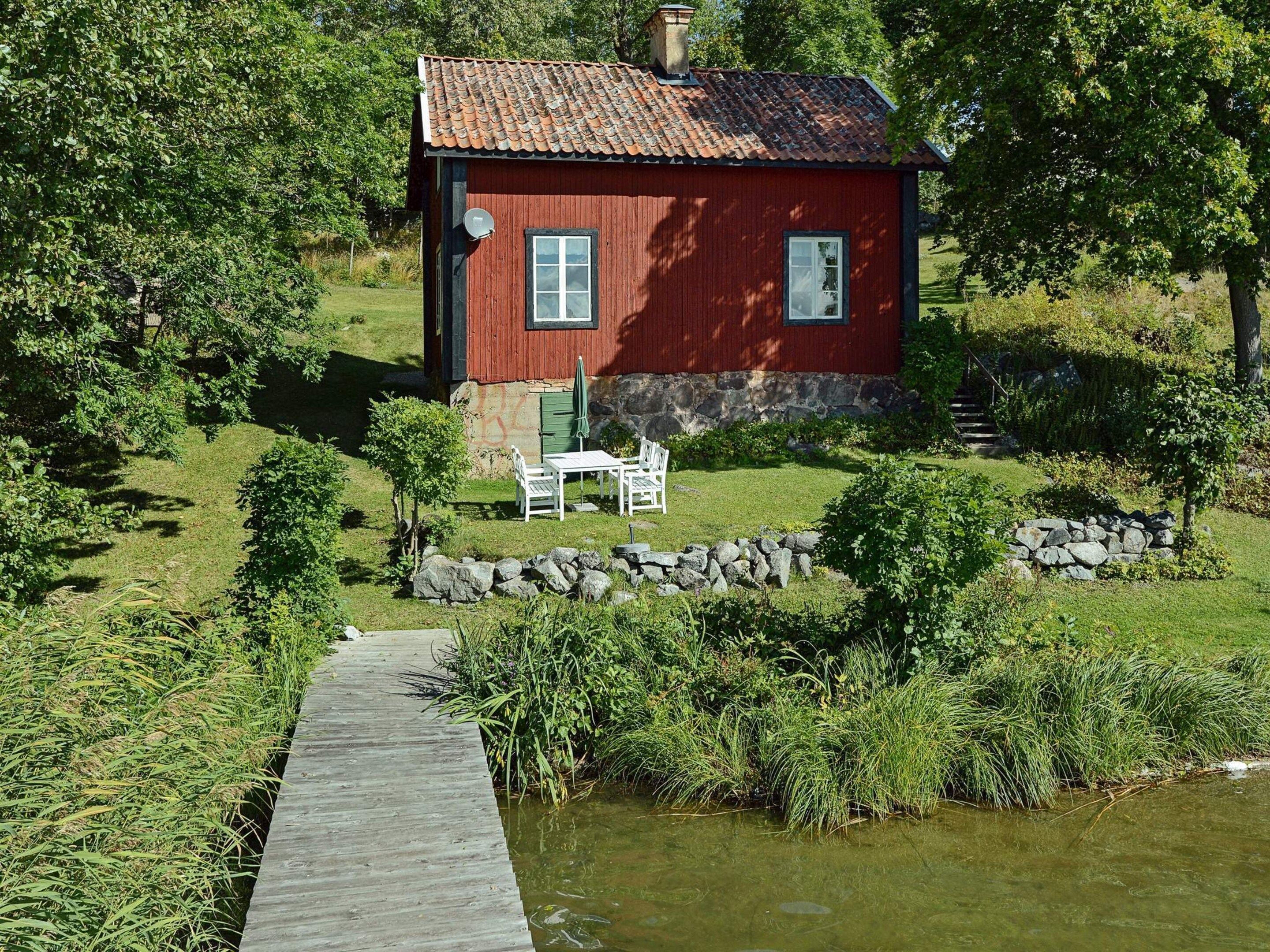 A red wooden house with a stone foundation, a small patio with a table and chairs, and a wooden dock leading to a body of water.