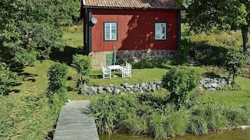 A red wooden house with a stone foundation, a small patio with a table and chairs, and a wooden dock leading to a body of water.