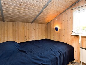 A room with wooden walls and a sloped ceiling, a bed with a dark cover, a wall-mounted light fixture, and a window with a white frame. - 4 Star Holiday Home in Stege (Stege)