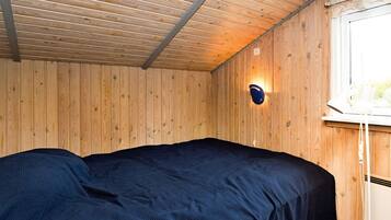 Una habitación con techo de madera, una cama individual con cubierta oscura, una lámpara de pared y un ventanal con una lámpara blanca en el alféizar.