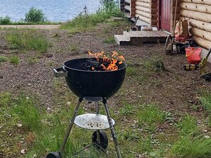 A black grill with flames, set on a metal stand, outdoors with grass and a wooden structure in the background. - Holiday Home in SÃ¤len (Lima)